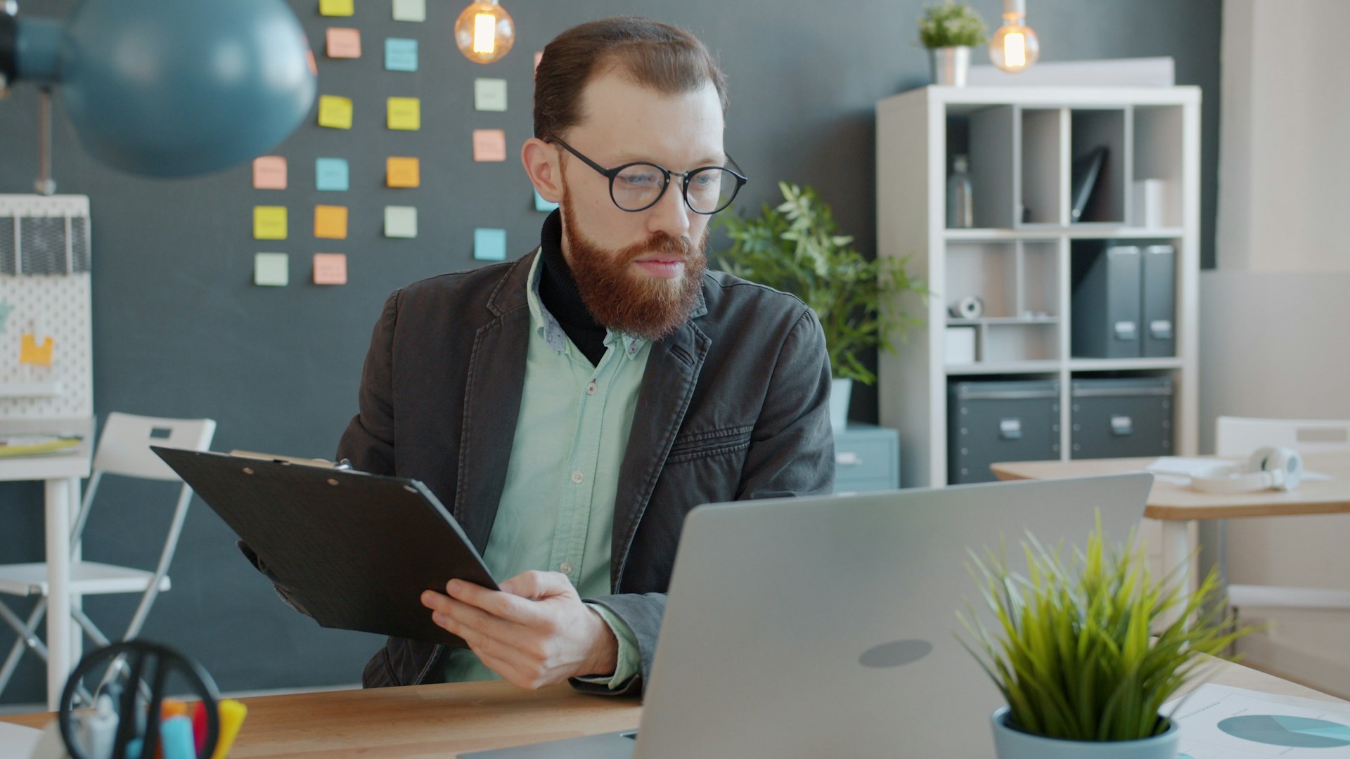 Man with beard working on laptop and clipboard.